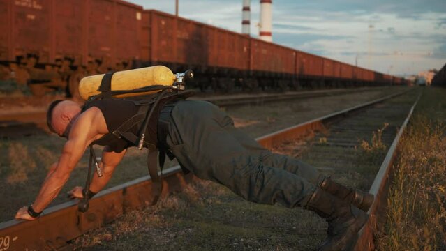 Young muscular man training at railway yard and moves on his hands and feet along the rails with drager ballon on his back
