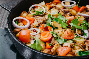 Fried potatoes with meat in a cast iron pan close-up with tomatoes, onions and parsley