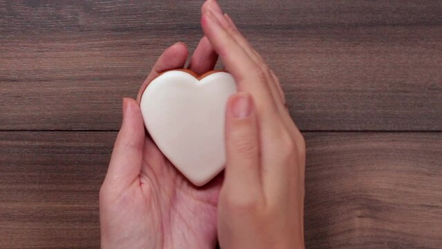 Female hands hold white big heart cookie and covering it with hand on wooden background. Love concept. Valentine day. Copy space