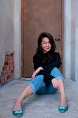 Beautiful southeast Asian woman wearing black sweater, jeans, and green shoes sits on the ground outside a stage door. Female gives a confident smile.