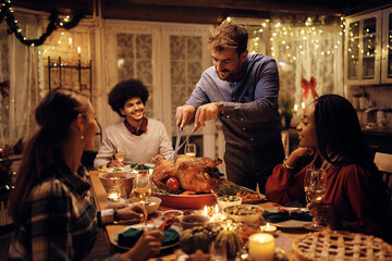 Happy man carving Thanksgiving turkey during dinner party with friends.