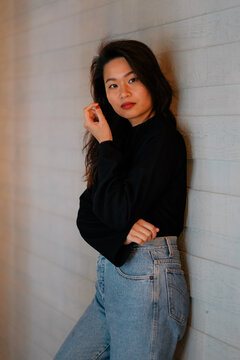 Southeast Asian woman wearing dark sweater and light jeans, leans on a wall and gives the camera a pointed look. Female has wavy hair and red nails.