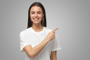 Smiling young woman in white t-shirt pointing right with index finger on gray background