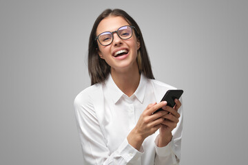 Positive laughing lady in glasses and white T-shirt holding smartphone, isolated on gray