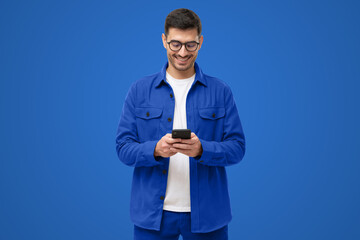 Young man in blue casual shirt looking at phone, standing on blue background
