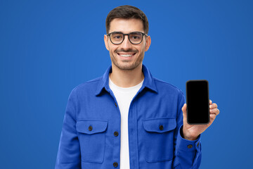 Young smiling man in casual shirt showing blank screen smartphone in hand, isolated on blue