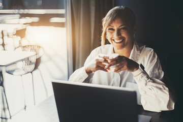 Portrait of a happy smiling Hispanic female entrepreneur in a white shirt, sitting with a cup of expresso coffee in her hands in front of her laptop next to the window of a cafe or a restaurant - obrazy, fototapety, plakaty