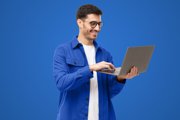 Young man wearing casual blue shirt, standing with opened laptop in hands, surfing online