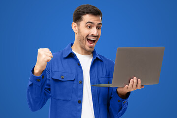 Excited man in blue shirt looking at laptop screen with WOW expression, isolated on blue