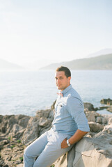 Groom sits on a rocky shore by the sea holding his hands in his pockets