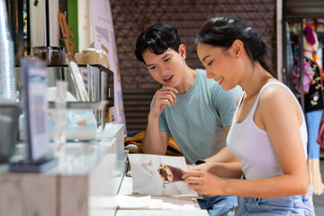 Young Asian couple enjoy and fun outdoor lifestyle shopping together at street market on summer holiday vacation. Happy man and woman buy ice coffee and soft drink at street market outdoor cafe.