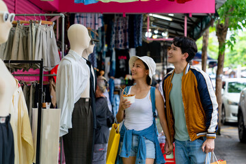 Young Asian couple enjoy and fun outdoor lifestyle shopping together at street market on summer holiday vacation. Happy man and woman choosing and buying fashion clothes together at clothing shop.