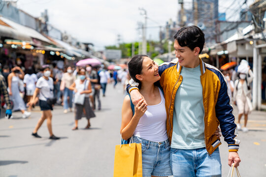 Attractive Asian couple enjoy and fun outdoor lifestyle shopping at street market while travel on summer holiday vacation. Happy man and woman couple holding shopping bag walking at weekend market