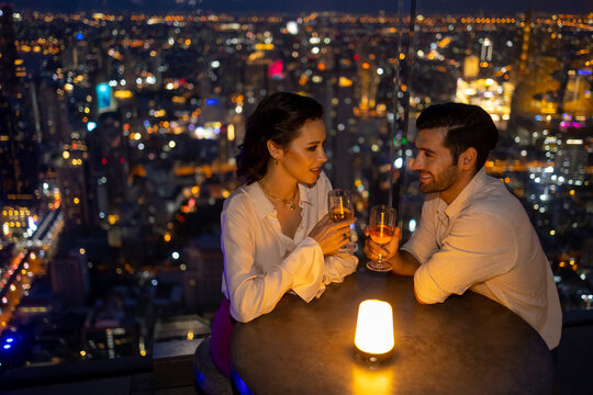 Caucasian Couple Celebrating Holiday Event At Luxury Skyscraper Rooftop Bar At Night. People Enjoy City Nightlife Having Dinner And Drinking Wine At Outdoor Rooftop Restaurant On Summer Vacation.