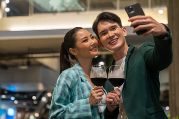 Asian couple celebrating holiday event at luxury restaurant at night. Man and woman couple using mobile phone taking selfie together while having dinner and drinking wine at bar on summer vacation