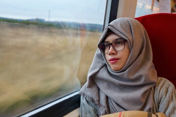Portrait of young and beautiful Asian Muslim woman wearing eyeglasses and hijab sitting alone against the window in a moving train. Looking out through window. Contemplating expression.