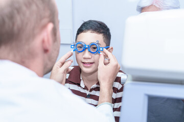 Smiling boy in glasses examining eyesight at a pediatric ophthalmologist  with grandma to support in modern clinic, doctors and ophthalmology clinic patients.
