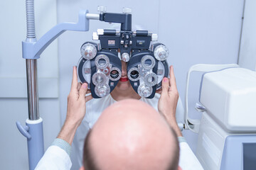 Senior Asian woman looking through optical phoropter during eye exam, diagnostic ophthalmology equipment, selective focus