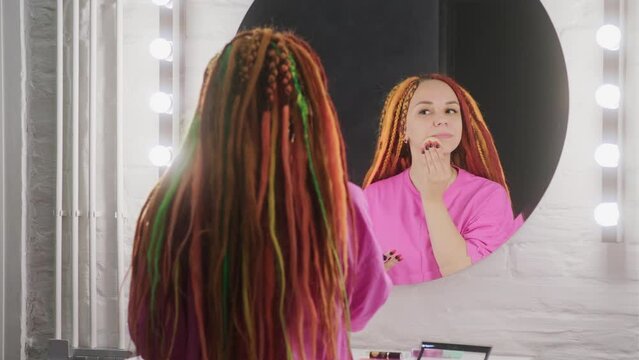 Young woman with dreadlocks looking in mirror, powdering her face. Pretty female with colourful multicoloured hairstyle preening in front of mirror, making make-up.