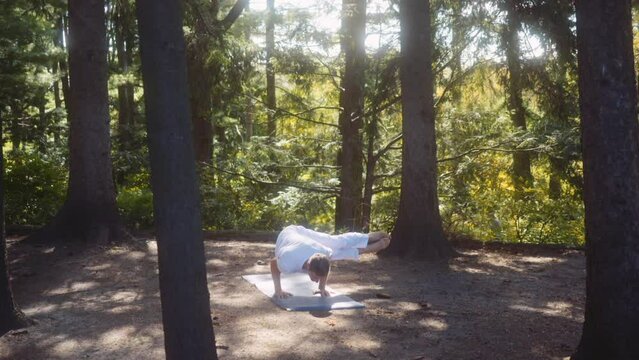 Caucasian adult man dressed in white walks to and gets on a yoga mat in a pine tree forest with great light. Camera handheld, wide shot. Side crow, parsva bakasana, advanced yogi.