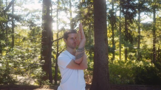 Close up shot caucasian adult man dressed in white holds a yoga pose in a pine tree forest.
a blend of tree pose and eagle pose: garudasana