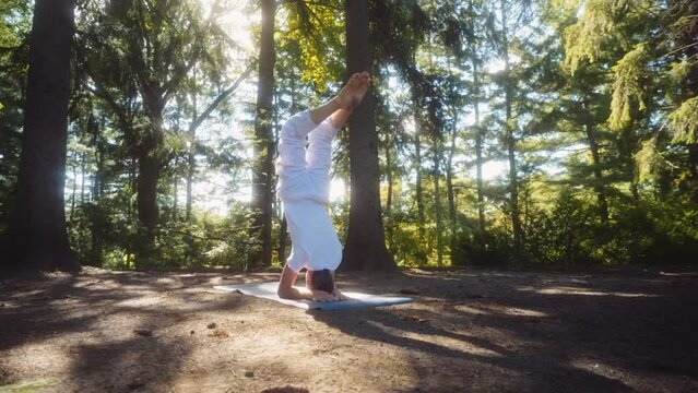 Caucasian adult man dressed in white holds then releases headstand pose on a yoga mat in a pine tree forest with great light. Camera handheld, wide shot, lens flare.