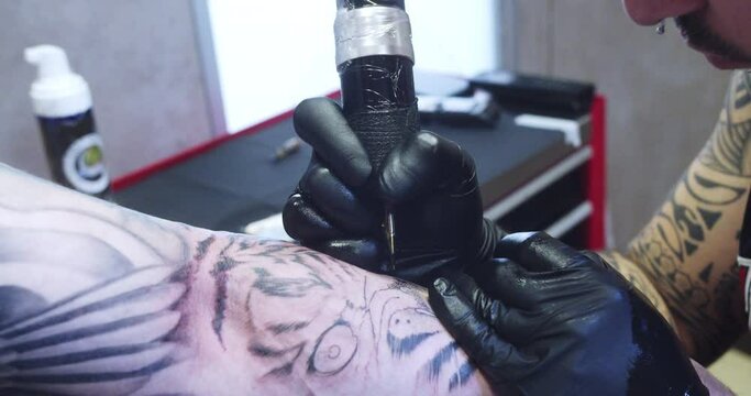 Side shot of a professional tattoo artist in the process of tattooing a man's arm inside a lighted studio.