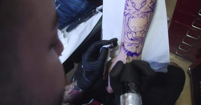 Moving close-up of the beginning of a tattoo on a man's arm. The studio is lit up and the tattoo trace is drawn in blue ink.