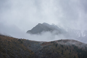 Dark atmospheric landscape with high mountain silhouettes in dense fog in rainy weather. Snowy rocky mountain top above hills in thick fog in dramatic overcast. Black rocks in low clouds during rain.