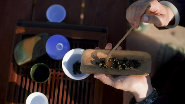 High angle view of cropped man pouring dry Oolong tea in cup. Traditional Chinese ceramic bowls on wooden chaban tray. He is preparing tea beverage during teatime on sunny day. 