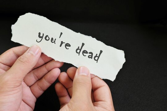 Death threat concept. Human hands holding a paper note with written word you're dead in dark black background.