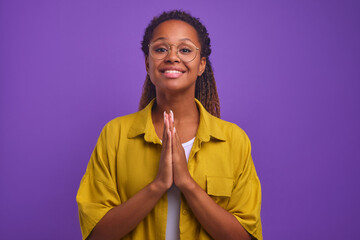 Young smiling African American woman looks at camera and makes gesture of prayer to ask for help or...