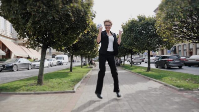 Full length woman waving dancing with dollars outside. Happy female entrepreneur businesswoman in formal suit keeping money stack at urban street. Smiling business woman holding us cash outdoors.