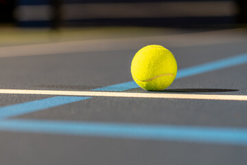 Photo taken in late evening under lights of a yellow tennis ball on blue tennis court with white...