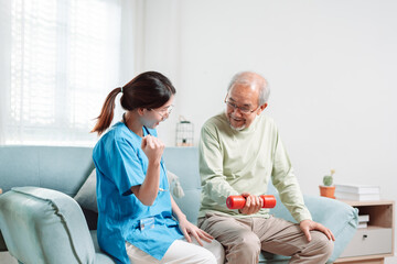 Asian Senior old man doing physiotherapist with support from caregiver. Mature male sitting on sofa in living room using dumbbell workout exercise. Healthcare medical concept.