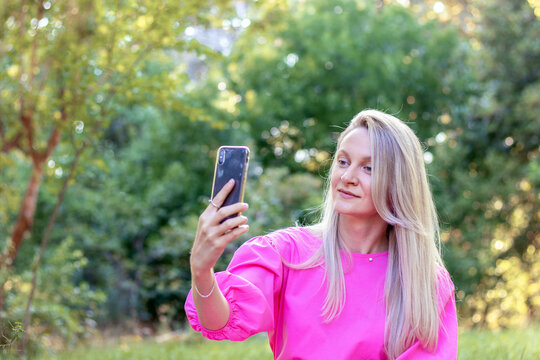 beautiful blonde girl woman in pink princess dress making taking selfie in middle of nature park green grass tree leaves.smiling happy female using technology smartphone harmony with nature.park back