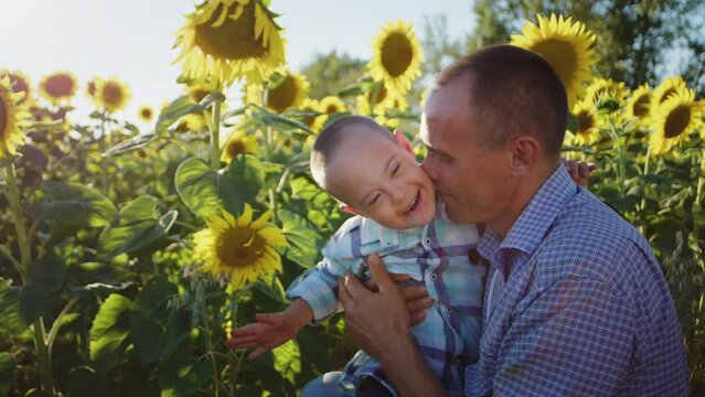 Man kisses little son with Down syndrome sitting among blooming sunflower field illuminated by bright sunlight. Caring father enjoys spending summer vacation with sick child in countryside closeup
