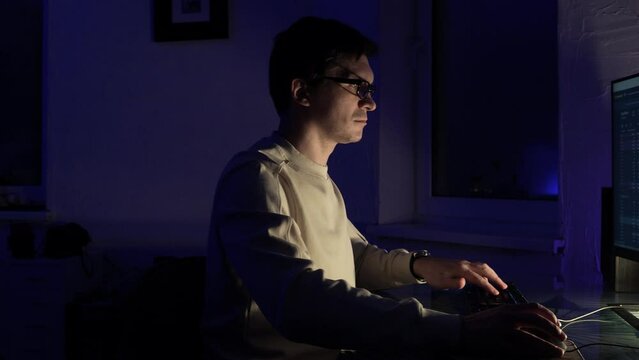 The male programmer works late. A guy with glasses is sitting in a dark office in front of a monitor and writing code.