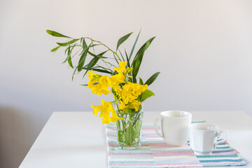 Shrub branch with Tecoma stans flowers in a glass beaker on a white table in the interior. A pink spring notepad, two coffee mugs and a geometric patterned napkin.