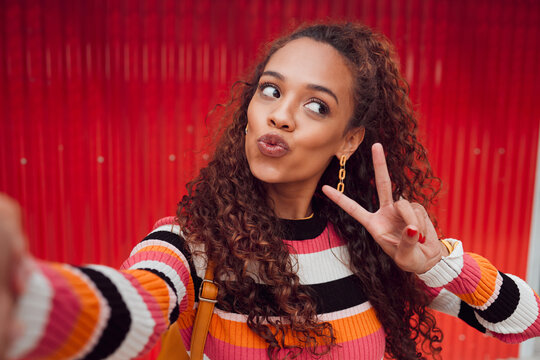 Fashion selfie, peace sign and happy woman against a red wall in city for fun travel holiday. Portrait urban girl show cool hand gesture and lips while traveling and sharing picture on social media