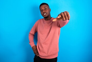 young handsome man wearing pink sweater over blue background pointing at camera with a satisfied,...