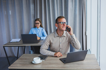 A handsome businessman is working during a video call on a laptop. The concept of a modern successful person. Young smiling male entrepreneur in glasses sits and works at a desk in an office space