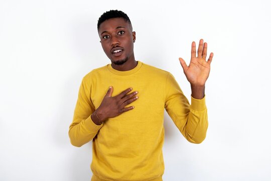 I swear, promise you not regret. Portrait of sincere young handsome man wearing yellow sweater over white raising one arm and hold hand on heart as give oath, telling truth, want you to believe.