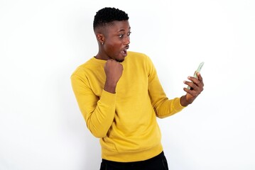 Happy cheerful young handsome man wearing yellow sweater over white background receiving good news via e-mail and celebrating success while standing and looking at mobile phone.