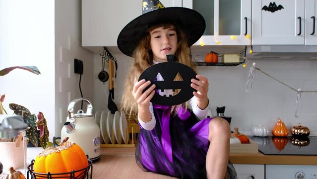 Child decorates the kitchen in home for Halloween. Girl in a witch costume plays with the decor for the holiday - bats, jack lantern, pumpkins. Autumn comfort in house, Scandi-style kitchen, loft