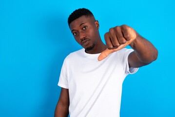 young handsome man wearing white T-shirt over blue background looking unhappy and angry showing rejection and negative with thumbs down gesture. Bad expression.