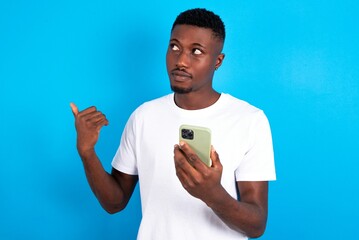 young handsome man wearing white T-shirt over blue background points thumb away and shows blank space aside, holds mobile phone for sending text messages.