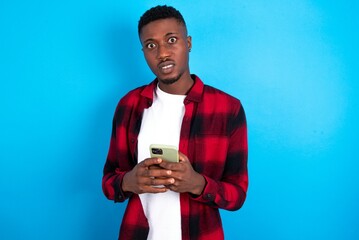 Photo of astonished crazy young handsome man wearing overshirt over blue background hold smartphone...