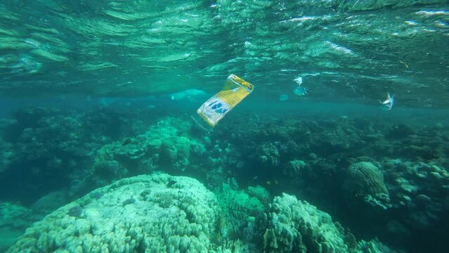 Slow motion, discarded plastic packaging slowly drifting under the surface of water nearby coral reef a tropical fish swimming around.  