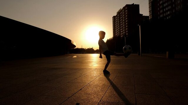 The silhouette of a little boy stuffing a ball in a city park in summer at sunset. The child is engaged in football.